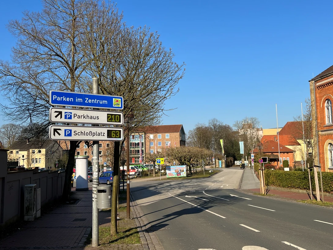 Straßenschild und Bäume entlang einer leeren Straße, die zu einem Parkplatz und Parkhaus führt.