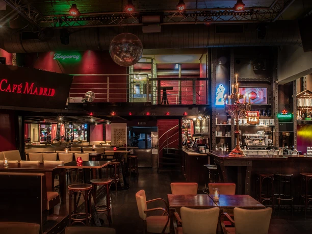 Cafe Madrid (c) Christoph Görres _ ORA GmbH (3).jpg In dem Raumausschnitt des Cafés befindet sich ein Teil der Theke sowie größere Sitzgruppen mit Stühlen und Bänken.