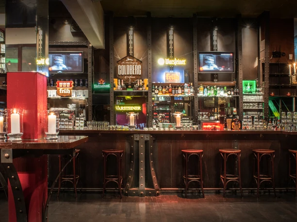 Cafe Madrid (c) Christoph Görres _ ORA GmbH (2).jpg Die Theke des Cafés befindet sich vor einem großen Flaschenregal und ist aus dunklem Holz. Davor stehen einige Barhocker.