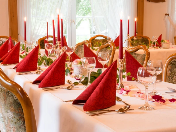 Festtafel Festlich gedeckte Tafel mit weißen Tischdecken, roten Stoffservietten und brennenden Stabkerzen in goldenen Haltern. Zwischen den Gedecken liegen grüne Blätter und Rosenblätter, umgeben von Kristallgläsern und silbernem Besteck, vor einem hellen Fenster mit lichtdurchfluteten Gardinen.