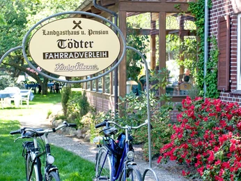 Garten 2.jpg Zwei Fahrräder stehen vor einem Schild mit der Aufschrift „Landgasthaus und Pension Tödter – Fahrradverleih“. Im Hintergrund sind ein Backsteingebäude mit begrüntem Wintergarten, blühende Büsche mit roten Blüten und Gartenmöbel auf einer Rasenfläche unter Bäumen zu sehen.
