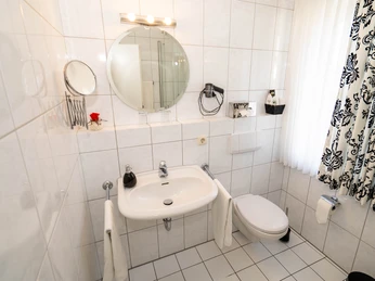 Badezimmer Hotel Papenbreer Kleines, hell gefliestes Badezimmer mit runden Spiegel, Waschbecken, WC und schwarz-weißen Vorhängen.