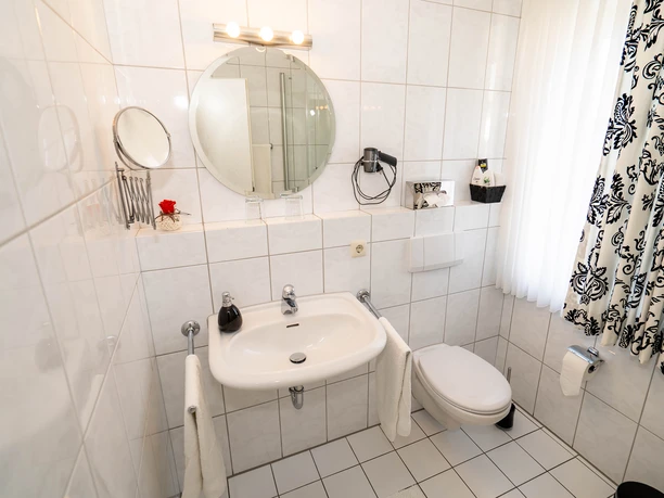 Badezimmer Hotel Papenbreer Kleines, hell gefliestes Badezimmer mit runden Spiegel, Waschbecken, WC und schwarz-weißen Vorhängen.