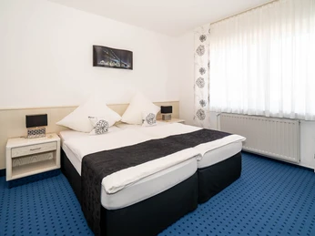 Hotelzimmer Hotel Papenbreer Helles Hotelzimmer mit zwei zusammengestellten Einzelbetten, blauen Teppichboden und hellen Vorhängen.