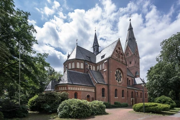 Eine neugotische Backsteinkirche mit spitzen Türmen, umgeben von Bäumen und blühenden Sträuchern.