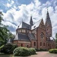Eine neugotische Backsteinkirche mit spitzen Türmen, umgeben von Bäumen und blühenden Sträuchern.