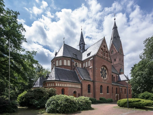 Eine neugotische Backsteinkirche mit spitzen Türmen, umgeben von Bäumen und blühenden Sträuchern.