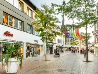 Eine belebte Einkaufsstraße mit Geschäften, Bäumen und bunt dekorierten Fahnen im Stadtzentrum.