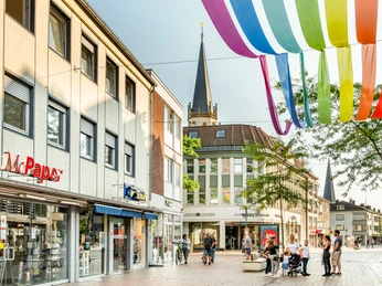 Geschäftsstraße in Wesel mit buntem Stoffdach und Kirchturm im Hintergrund, Menschen flanieren.