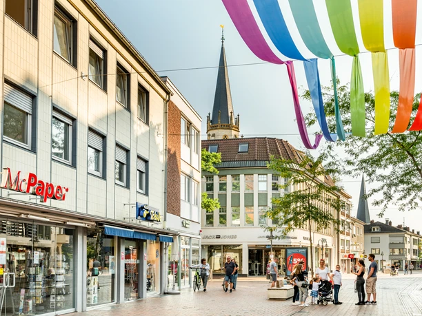 Regenbogenhimmel Innenstadt Gütersloh Geschäftsstraße in Wesel mit buntem Stoffdach und Kirchturm im Hintergrund, Menschen flanieren.