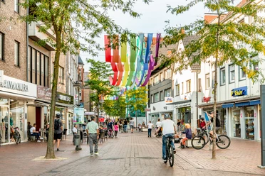 Eine belebte Fußgängerzone mit Geschäften, bunten Bannern und Menschen beim Einkaufen und Radfahren.