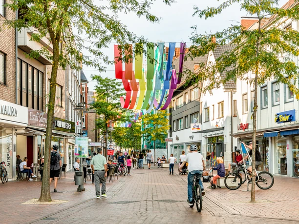 Eine belebte Fußgängerzone mit Geschäften, bunten Bannern und Menschen beim Einkaufen und Radfahren.
