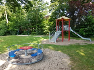 Spielplatz mit Rutsche, Schaukel und Karussell, umgeben von grünen Bäumen auf einer Wiese.