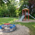 Spielplatz mit Rutsche, Schaukel und Karussell, umgeben von grünen Bäumen auf einer Wiese.