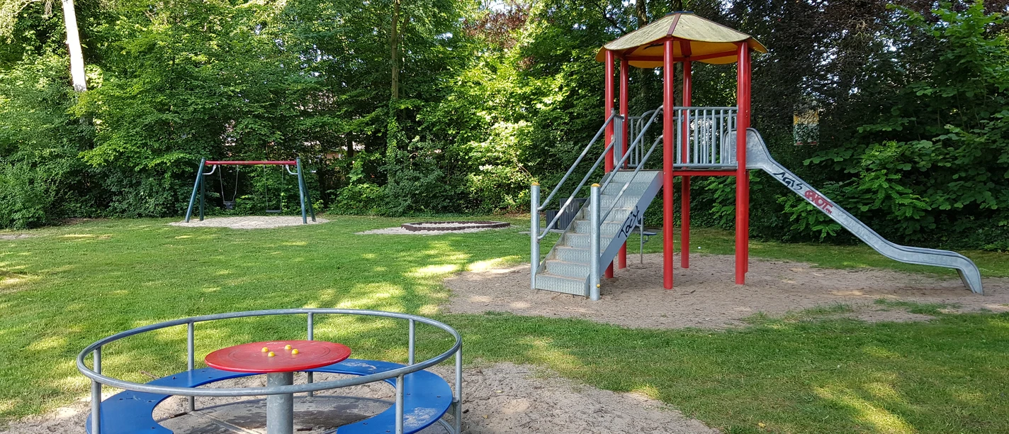 Spielplatz Elsternweg Spielplatz mit Rutsche, Schaukel und Karussell, umgeben von grünen Bäumen auf einer Wiese.