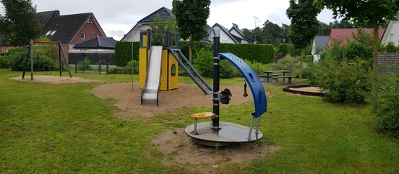 Ein moderner Spielplatz mit Rutsche, Schaukeln und Drehscheibe, umgeben von Häusern und Grün.