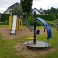 Ein moderner Spielplatz mit Rutsche, Schaukeln und Drehscheibe, umgeben von Häusern und Grün.