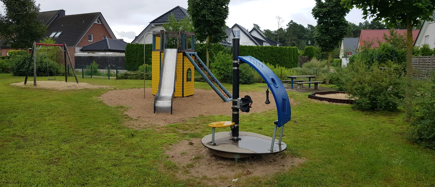 Ein moderner Spielplatz mit Rutsche, Schaukeln und Drehscheibe, umgeben von Häusern und Grün.