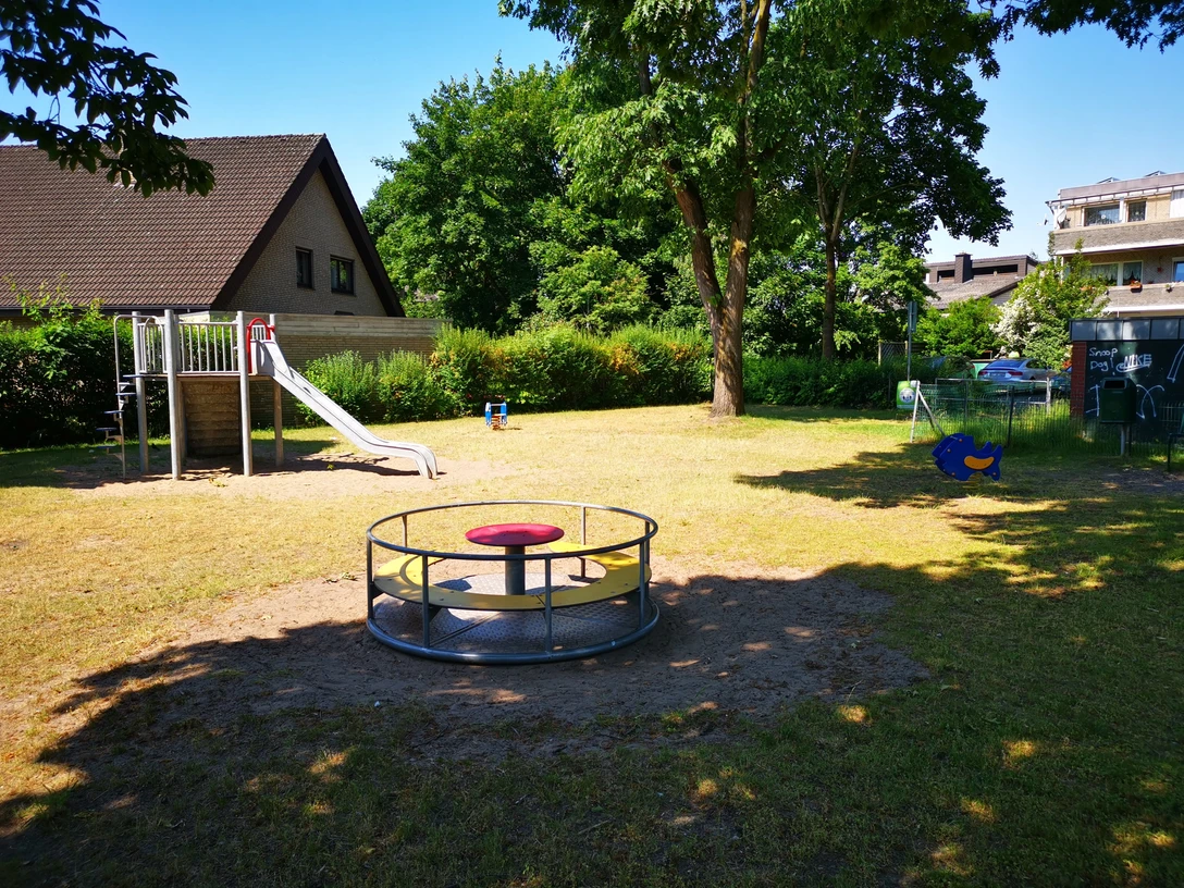 Ein kleiner Spielplatz mit Rutsche, Kletterturm, Wippe und Karussell, umgeben von Bäumen und Wiese.