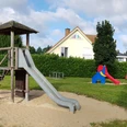 Spielplatz mit Holz-Klettergerüst, Rutsche, Schaukeln und buntem Spielhäuschen auf Sandfläche.