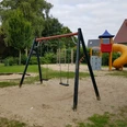 Ein Spielplatz mit einer Schaukel, Klettergerüst, Rutsche und Sandboden, umgeben von Bäumen und Büschen.