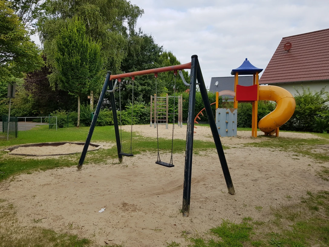 Spielplatz Köldingsweg Ein Spielplatz mit einer Schaukel, Klettergerüst, Rutsche und Sandboden, umgeben von Bäumen und Büschen.