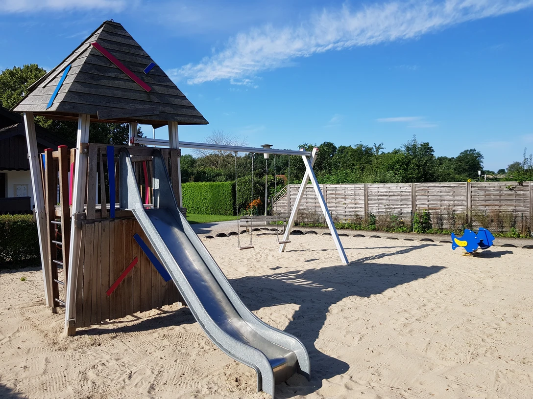 Spielplatz mit Holzturm und Rutsche auf Sandfläche, umgeben von Zaun und grünem Bewuchs.
