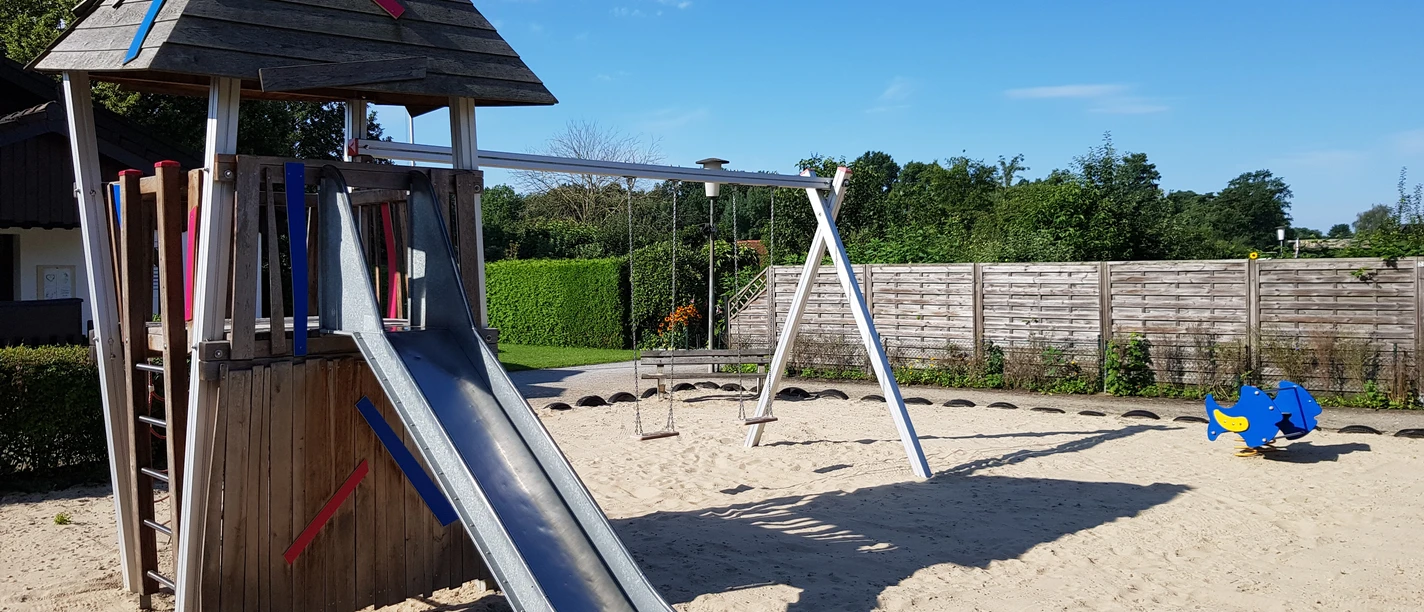 Spielplatz mit Holzturm und Rutsche auf Sandfläche, umgeben von Zaun und grünem Bewuchs.