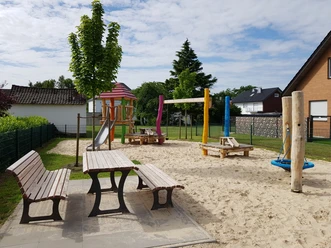 Spielplatz mit buntem Klettergerüst, Rutsche, Schaukeln und Picknicktischen auf hellem Sanduntergrund.
