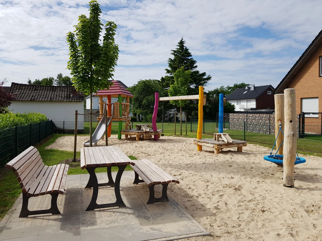 Spielplatz Lupinenweg Spielplatz mit buntem Klettergerüst, Rutsche, Schaukeln und Picknicktischen auf hellem Sanduntergrund.