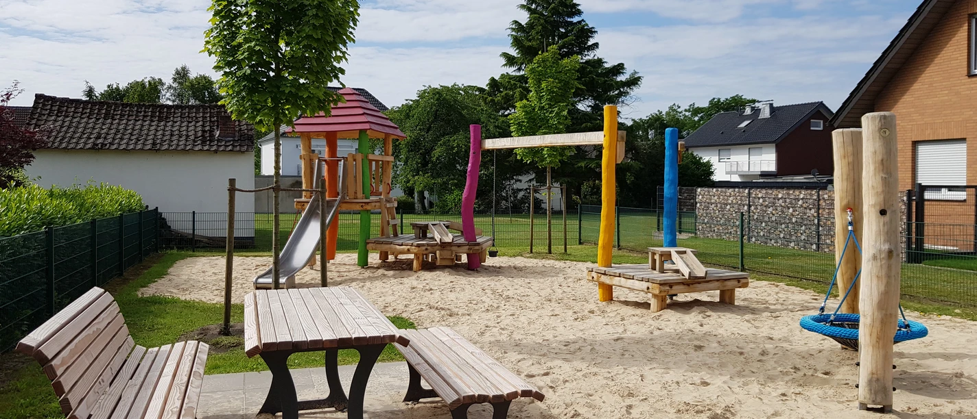 Spielplatz Lupinenweg Spielplatz mit buntem Klettergerüst, Rutsche, Schaukeln und Picknicktischen auf hellem Sanduntergrund.