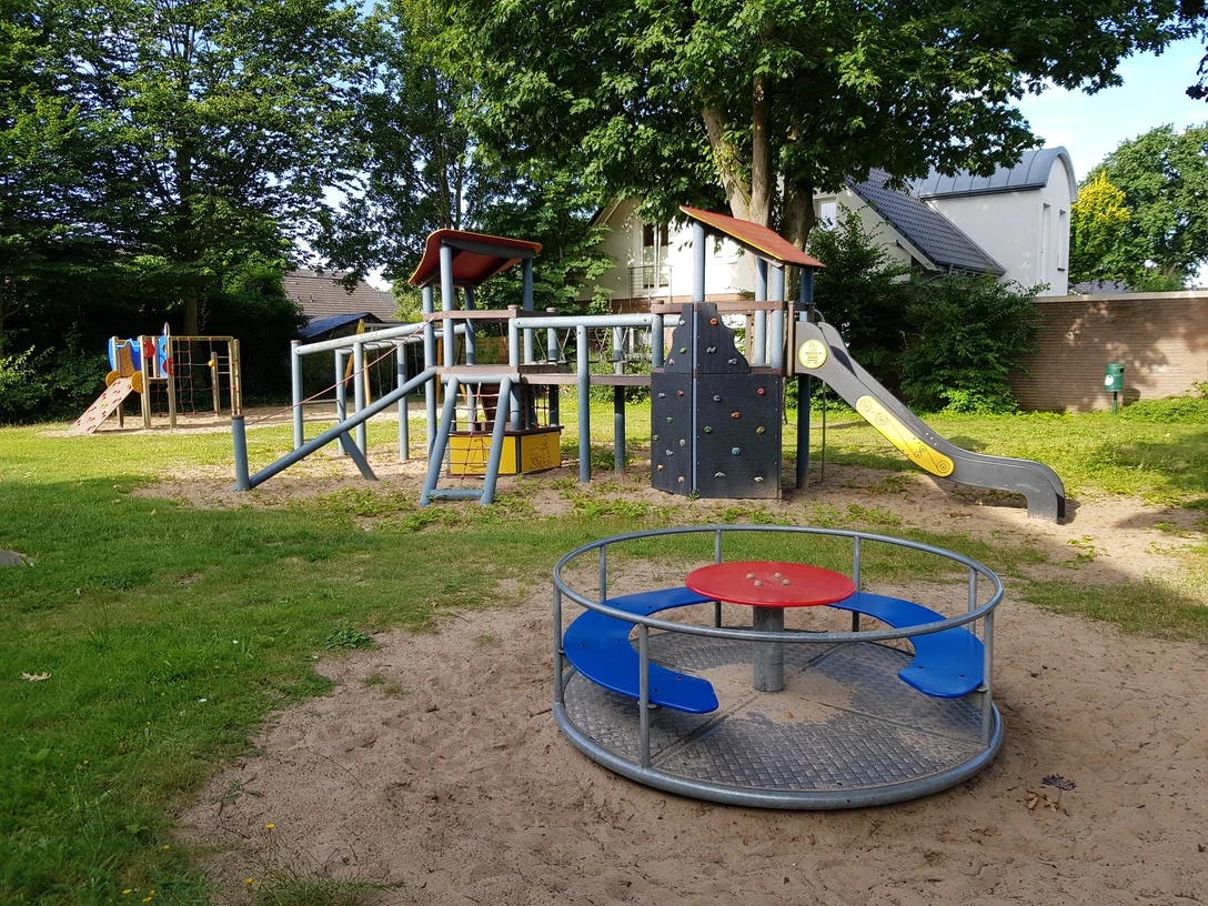 Kinderspielplatz mit Sandboden, Rutsche, Klettergerüst und Karussell, umgeben von grünen Bäumen.
