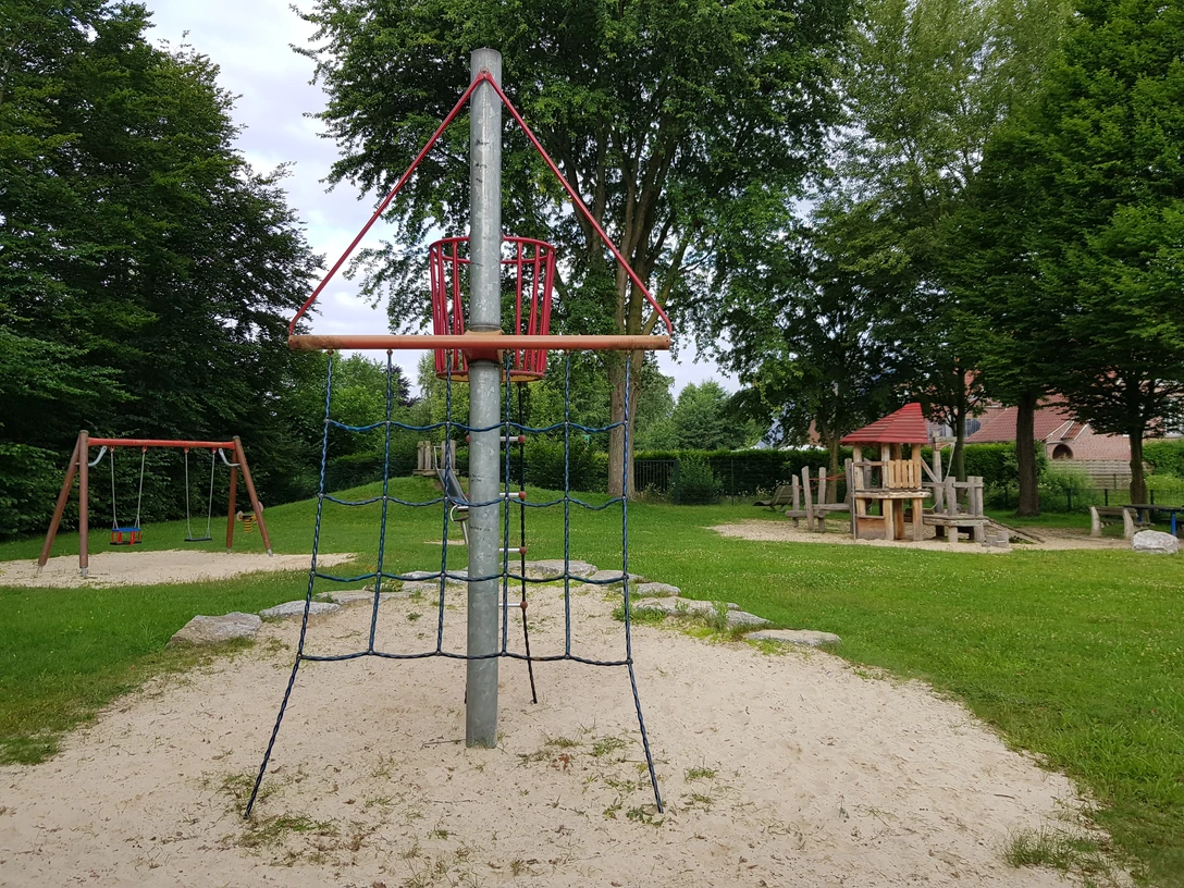 Spielplatz mit Klettergerüst, Schaukel und Balancierbereich, umgeben von grünem Rasen und Bäumen.