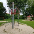 Spielplatz mit Klettergerüst, Schaukel und Balancierbereich, umgeben von grünem Rasen und Bäumen.