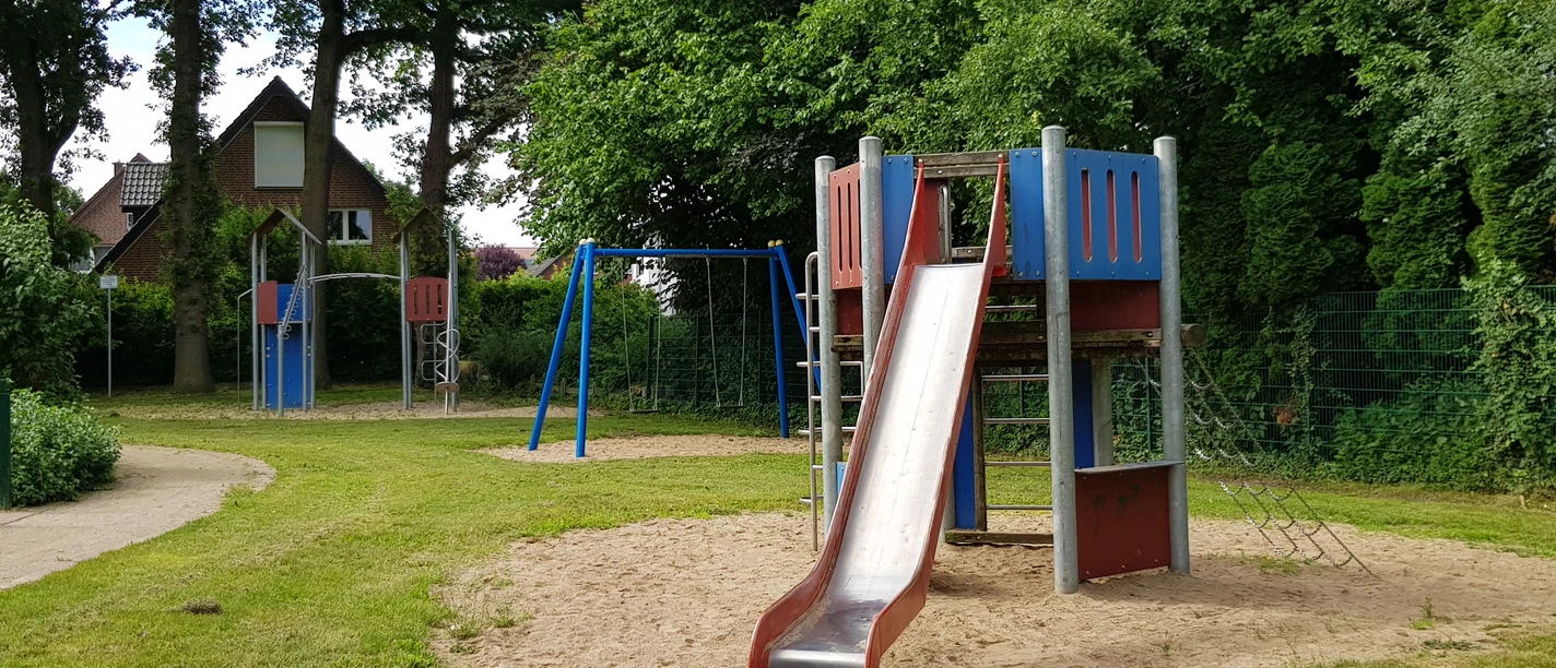 Kinderspielplatz mit Rutsche, Klettergerüst und Schaukeln, umgeben von grünen Bäumen und Rasenflächen.