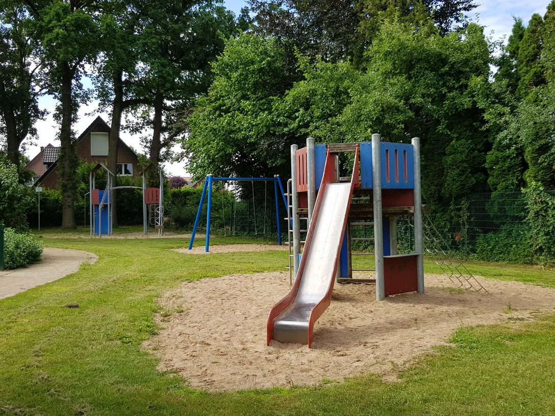 Spielplatz Rebhuhnweg Kinderspielplatz mit Rutsche, Klettergerüst und Schaukeln, umgeben von grünen Bäumen und Rasenflächen.