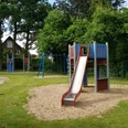 Spielplatz Rebhuhnweg Kinderspielplatz mit Rutsche, Klettergerüst und Schaukeln, umgeben von grünen Bäumen und Rasenflächen.