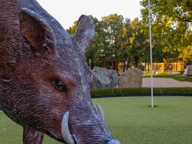 Minigolfanlage-lueneburger-salzsau-foto_ccbysa_foto©adventure-schnucken-golf-min.jpg Ein Wildschwein-Modell thront auf einer grünen Minigolfbahn, flankiert von einer Fahne und umgeben von beruhigender Natur.