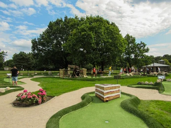 Minigolfanlage-schneverdingen_ccbysa_foto©adventure-schnucken-golf-min.jpg Eine grüne Minigolfanlage in Schneverdingen mit Menschen, die auf den Bahnen spielen. Im Hintergrund stehen Bäume, ringsum sind gepflegte Rasenflächen und Blumen zu sehen. Der Himmel ist teils bedeckt, aber freundlich.