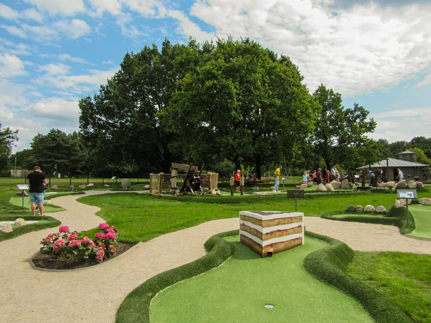 Minigolfanlage-schneverdingen_ccbysa_foto©adventure-schnucken-golf-min.jpg Eine grüne Minigolfanlage in Schneverdingen mit Menschen, die auf den Bahnen spielen. Im Hintergrund stehen Bäume, ringsum sind gepflegte Rasenflächen und Blumen zu sehen. Der Himmel ist teils bedeckt, aber freundlich.