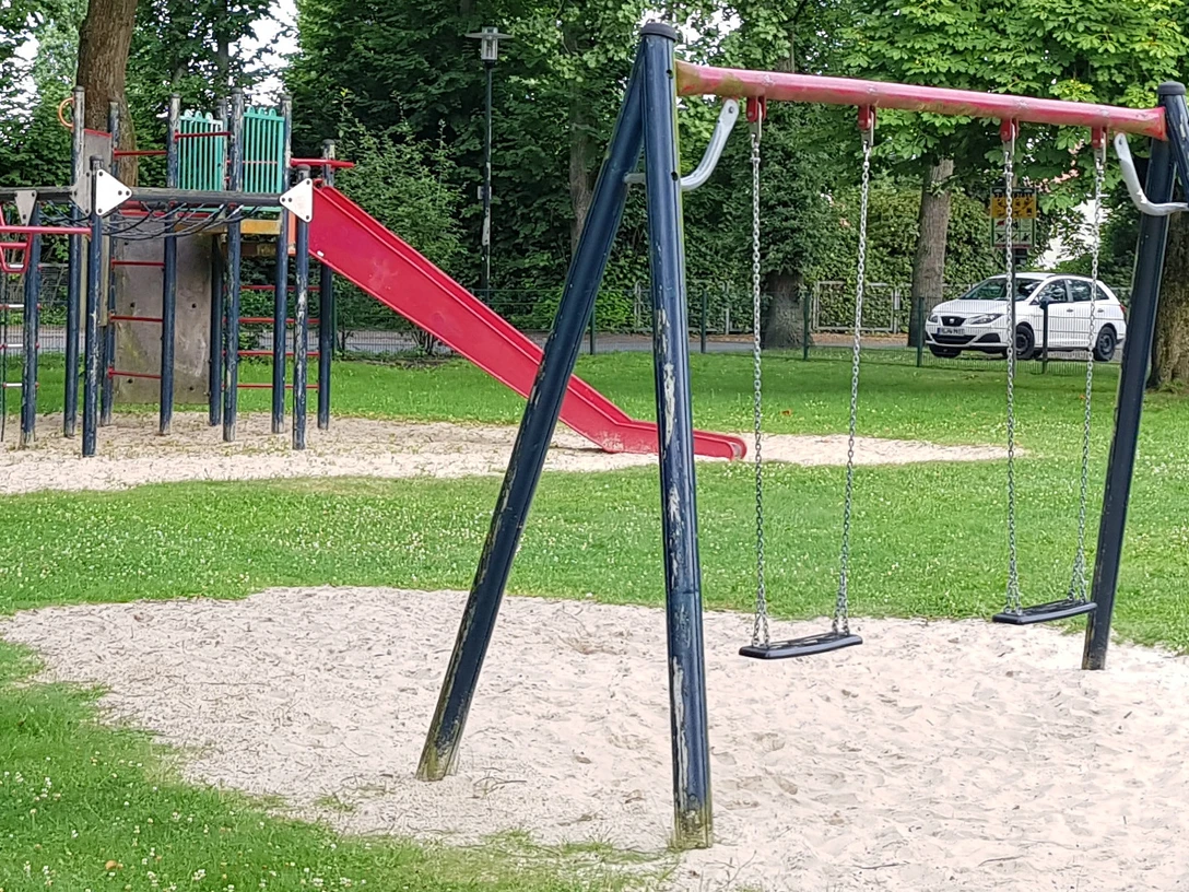 Spielplatz mit roter Rutsche, schwarzen Schaukeln und Klettergerüst auf grünem Rasen mit Bäumen.