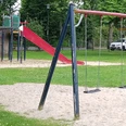 Spielplatz mit roter Rutsche, schwarzen Schaukeln und Klettergerüst auf grünem Rasen mit Bäumen.