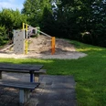 Ein Spielplatz mit Kletterwand, Holzklötzen und Sandfläche, umgeben von grüner Wiese und Bäumen.