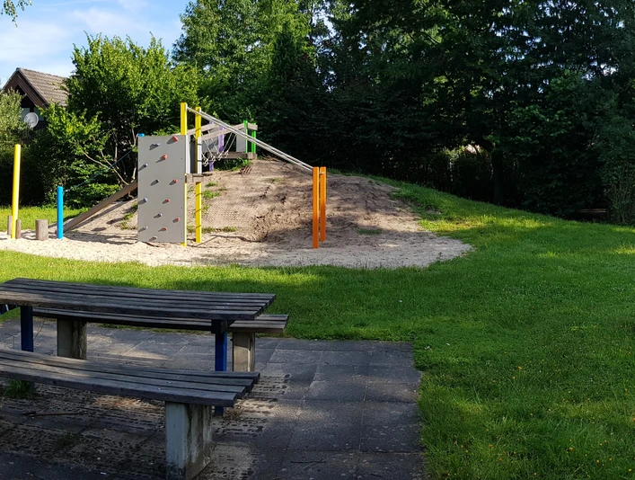 Ein Spielplatz mit Kletterwand, Holzklötzen und Sandfläche, umgeben von grüner Wiese und Bäumen.