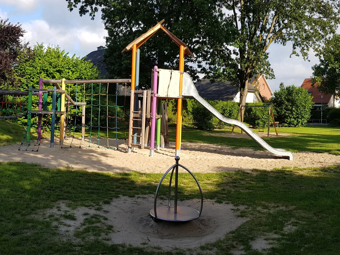 Spielplatz Westfalenweg Ein Spielplatz mit Rutsche und Klettergerät umgeben von Wiesen in einer sonnigen, grünen Umgebung.