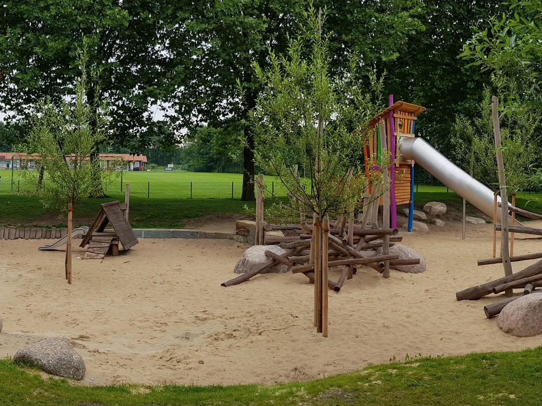 Ein großzügiger Spielplatz mit Rutsche, Sandfläche und Holzklettergerüst, umgeben von grünen Bäumen.