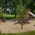 Ein großzügiger Spielplatz mit Rutsche, Sandfläche und Holzklettergerüst, umgeben von grünen Bäumen.