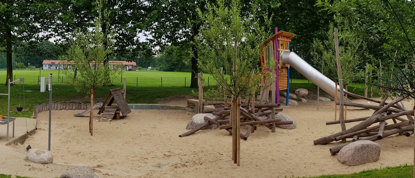 Ein großzügiger Spielplatz mit Rutsche, Sandfläche und Holzklettergerüst, umgeben von grünen Bäumen.