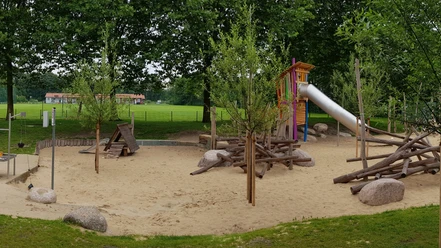 Spielplatz Trakehner Straße Ein großzügiger Spielplatz mit Rutsche, Sandfläche und Holzklettergerüst, umgeben von grünen Bäumen.