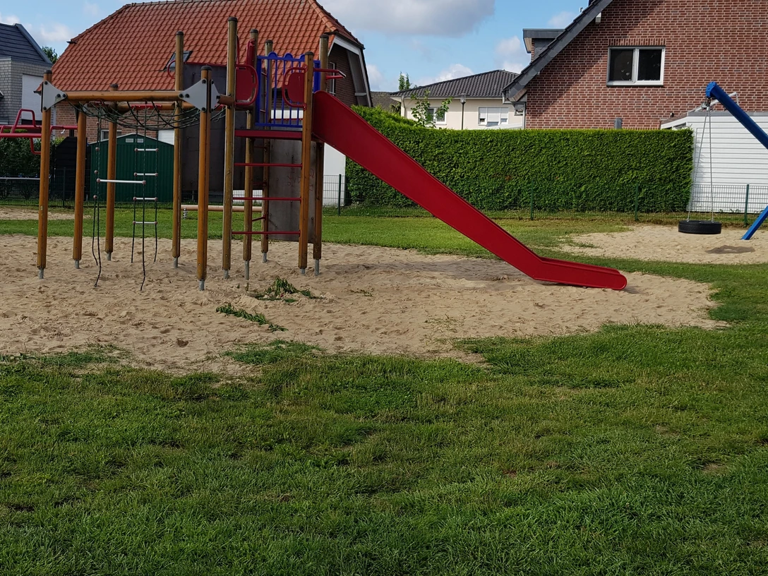 Ein Spielplatz mit Klettergerüst, Rutsche und Wippe, umgeben von Rasen und modernen Wohnhäusern.